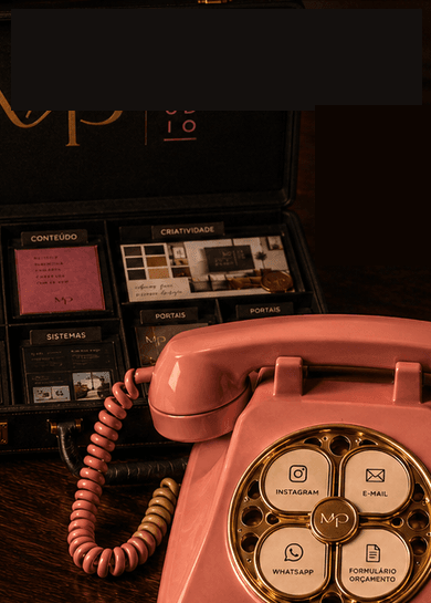 Telefone rosa vintage sobre a mesa quente da MPStudio, ao lado da maleta aberta, luminária, martini, caderno de couro e caneta